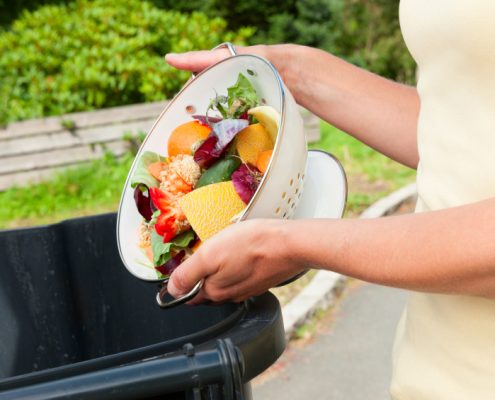 Unrecognizable woman emptying a collander of fruit and vegetable waste into a black plastic bin.